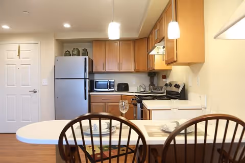 A small kitchen area with wooden cabinets, a stainless steel refrigerator, microwave, toaster, and stove. In the foreground, there is a white countertop with two place settings and two wooden chairs. The kitchen is well-lit with ceiling lights and pendant lights above the counter.