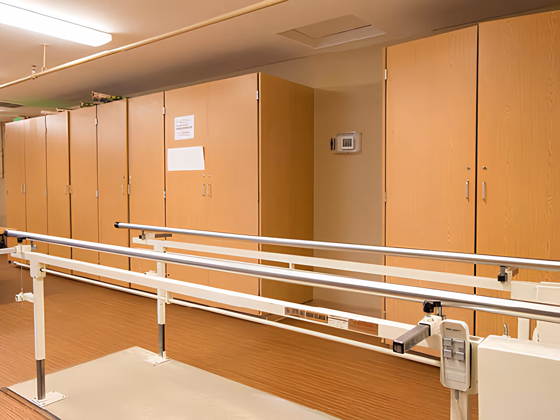 Parallel walking rails for physical therapy in a room with wood-paneled storage cabinets along the wall.