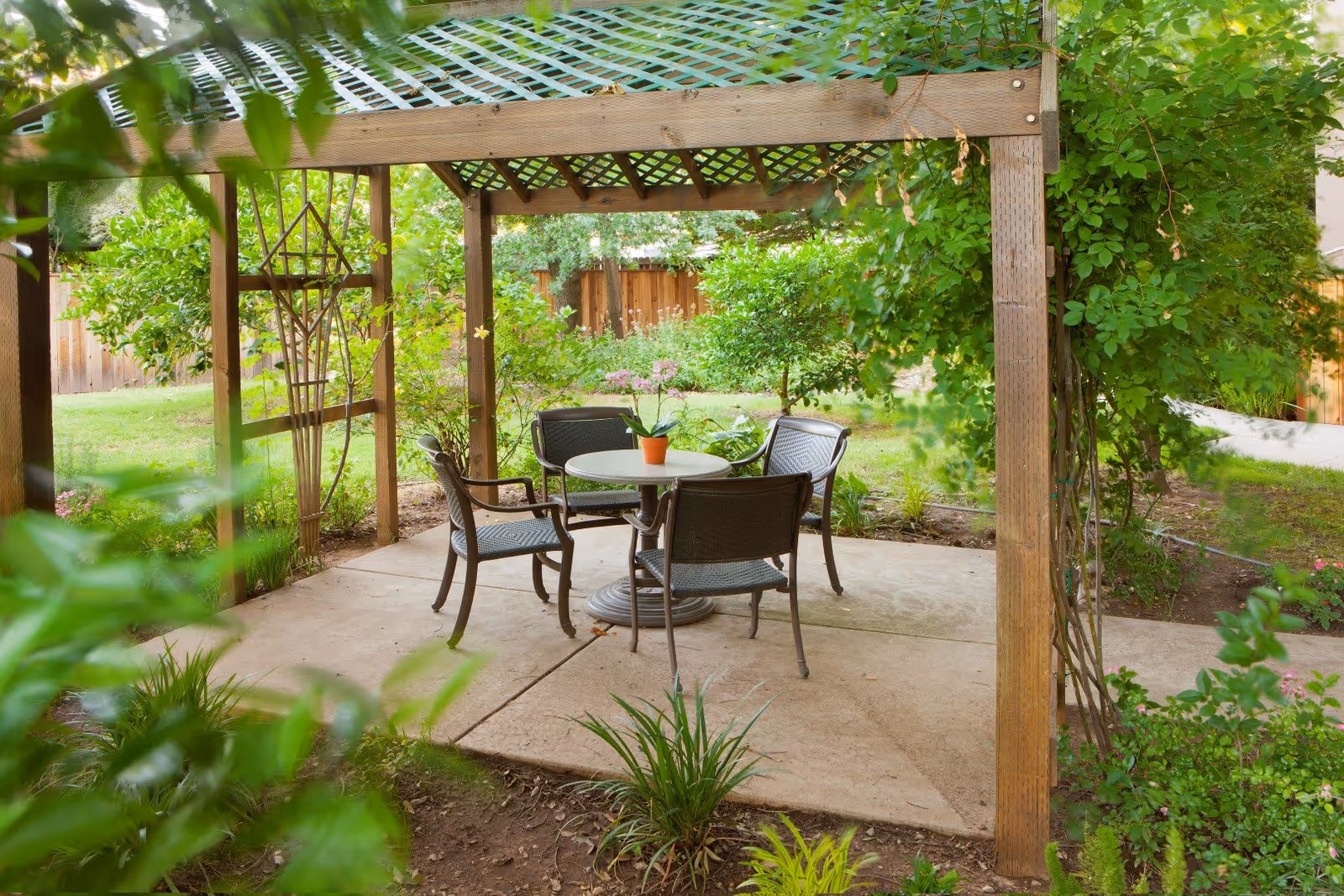 A wooden pergola in a garden setting with a round table and four chairs underneath. The table has a small potted plant with pink flowers. The surrounding area is lush with green plants, bushes, and trees, and a wooden fence is visible in the background.