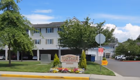 Exterior view of The Vintage at Mount Vernon senior living facility showing a multi-story building with covered parking underneath, a landscaped area with trees and flowers, and a sign that reads 'Vintage at Mount Vernon'. There is a street with a stop sign and a leasing office sign visible.