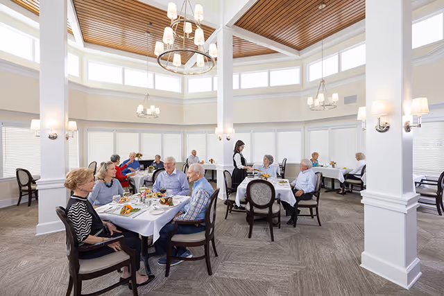 Elderly residents seated at white-clothed tables in a bright, high-ceiling dining room with a server attending to them.