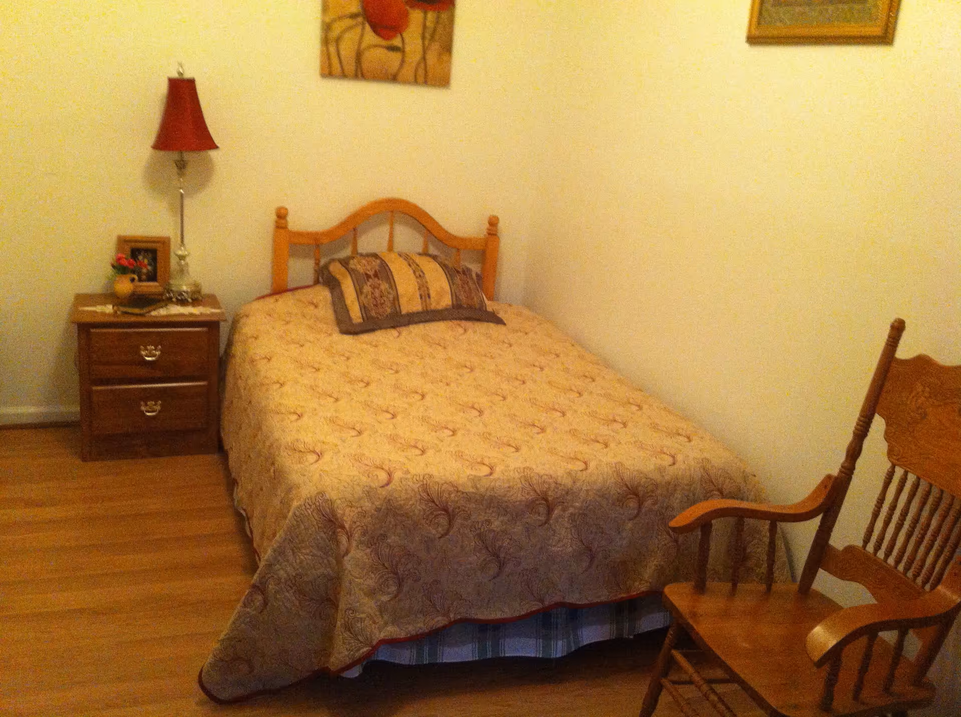 A small bedroom with a single bed covered in a beige patterned bedspread and a matching pillow. Next to the bed is a wooden nightstand with two drawers, a red lampshade on a silver lamp, a small framed photo, and a small potted plant. There is a wooden rocking chair to the right of the bed. The walls are plain and light-colored, with two framed pictures hanging above the bed.