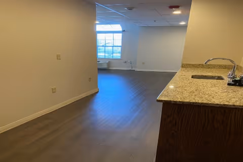 Interior view of an empty room with wood flooring, a large window letting in natural light, and a kitchen counter with a sink on the right side.