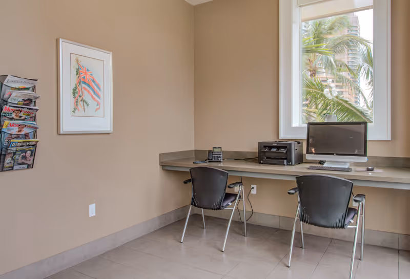 A small office or business center area with a long counter against the wall holding a desktop computer, printer, and a telephone. Two black chairs are placed in front of the counter. A window shows palm trees outside. On the left wall, there is a framed picture and a wall-mounted magazine rack with various magazines.