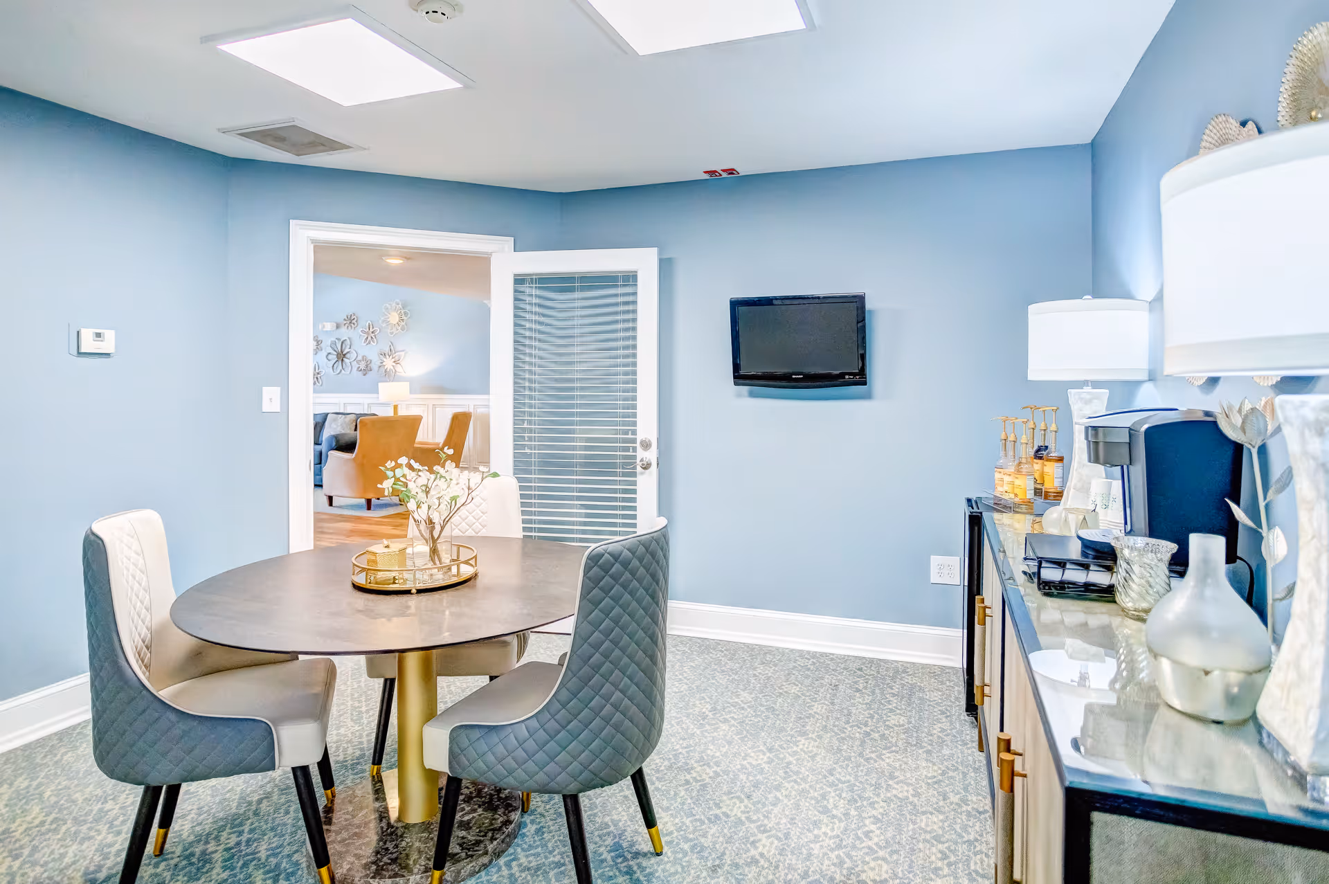 A small dining area with a round table and four cushioned chairs with quilted backs. The walls are painted light blue, and there is a flat-screen TV mounted on the wall. A glass door leads to another room with additional seating. On the right side, there is a countertop with a coffee maker, decorative items, and a lamp.
