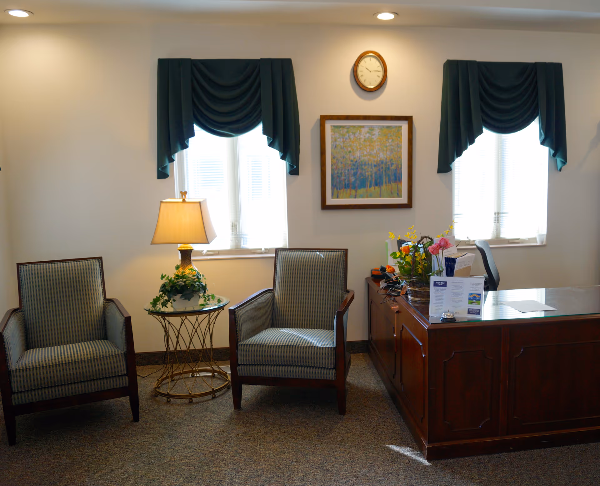 A cozy office or reception area with two upholstered armchairs separated by a small round glass table with a lamp and a plant. Behind the chairs are two windows with dark green drapes. A wooden desk with flowers, papers, and a chair is positioned to the right. A framed painting and a clock hang on the wall between the windows.