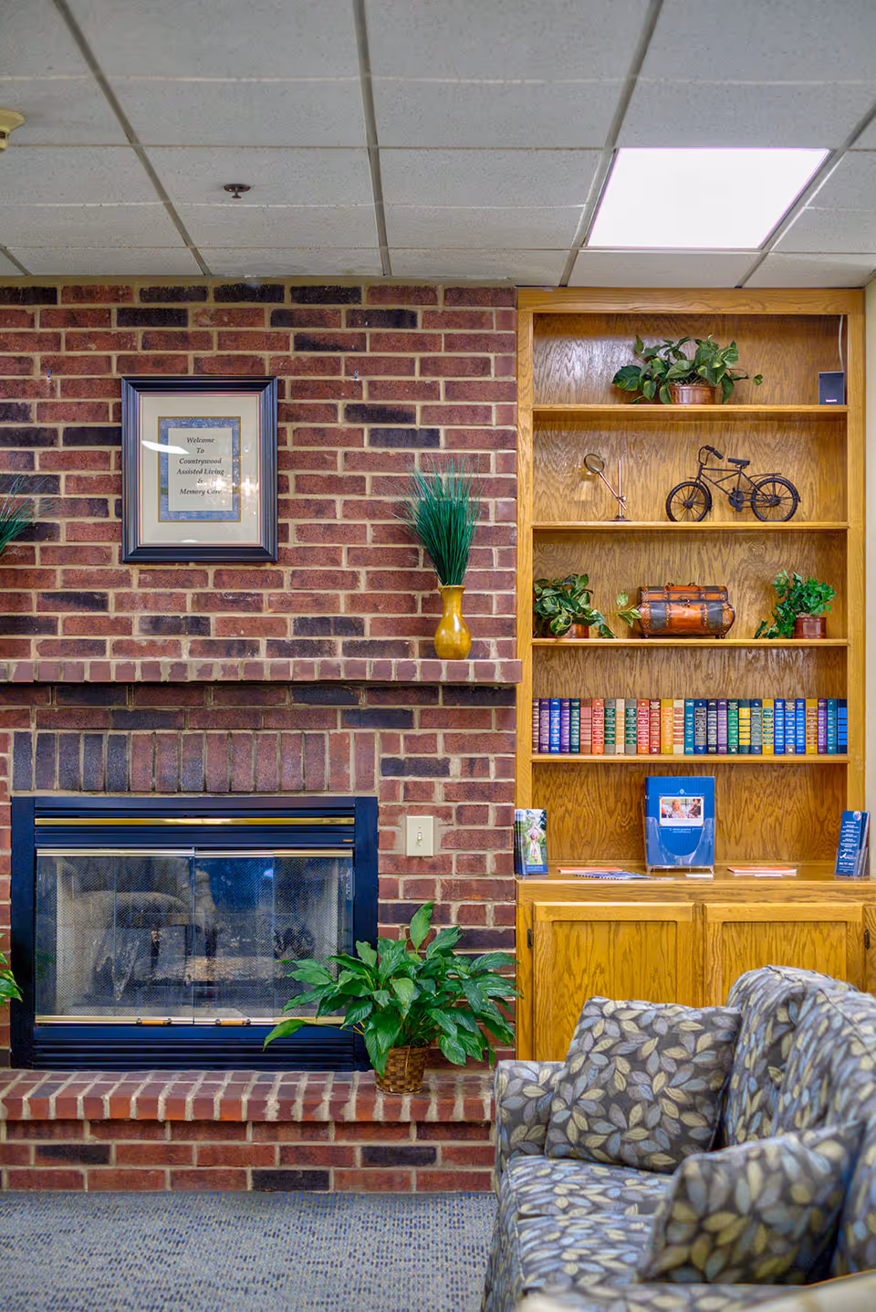 Interior view of a cozy living room area featuring a brick fireplace with a framed sign above it, a wooden bookshelf filled with books and decorative items, and a patterned armchair in the foreground.