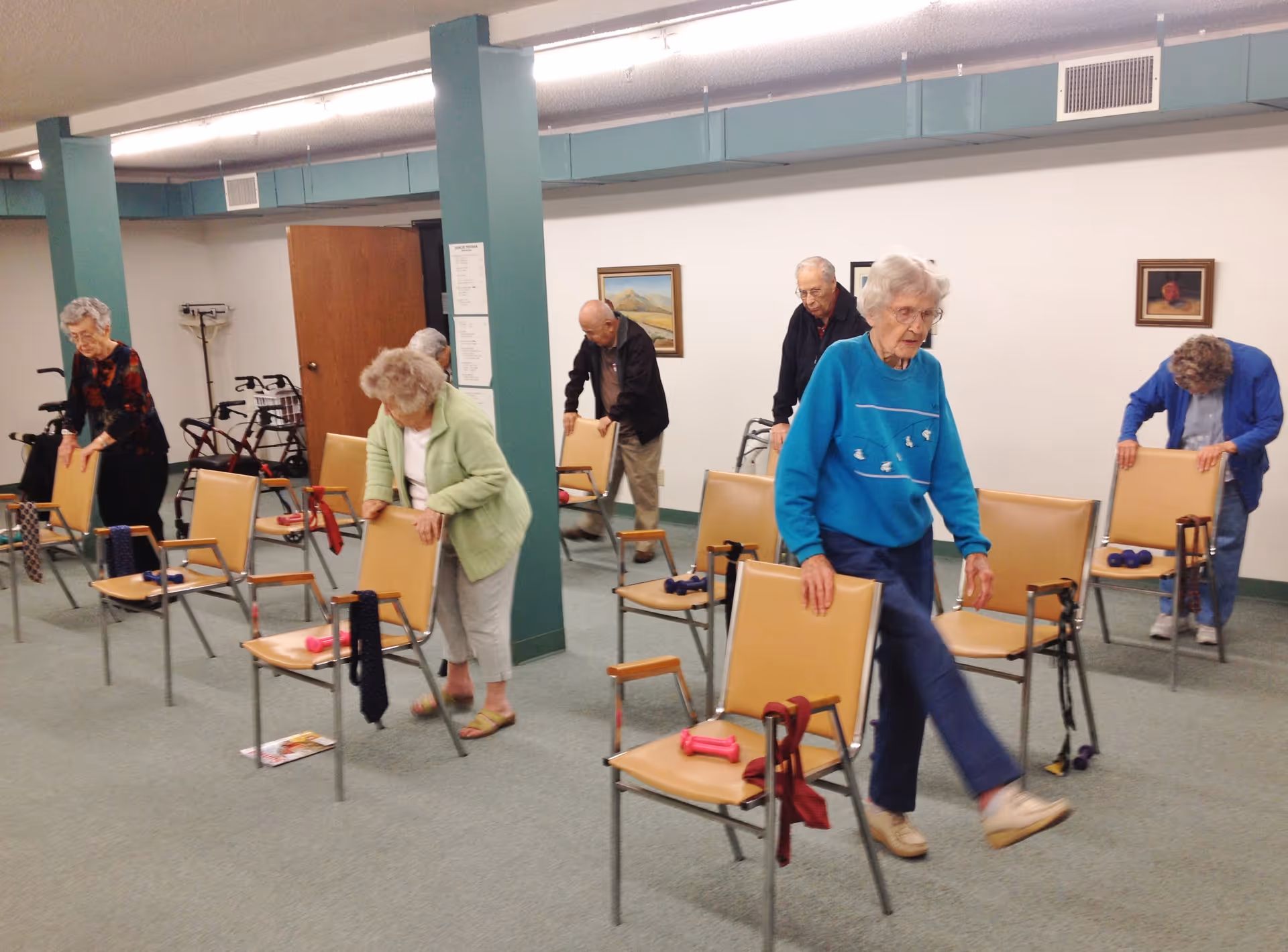 A group of elderly individuals participating in a seated exercise class in a community room. Each person is using a chair for support while performing leg lifts. The room has green pillars, beige chairs, and framed artwork on the walls.