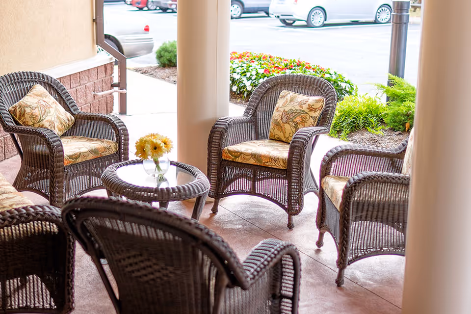 Wicker chairs with floral cushions arranged around a small glass-top table holding a vase of yellow flowers on a covered outdoor patio near the facility entrance.