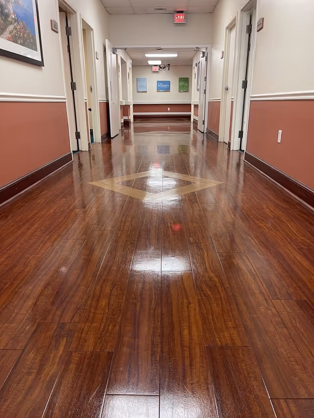 A clean, polished wooden floor hallway in a senior living facility with several open doors on both sides. The walls are painted white on the upper half and a reddish-brown on the lower half, separated by a white chair rail. There is a framed picture on the left wall and three colorful posters on the far wall. The ceiling has fluorescent lights and an exit sign is visible at the end of the hallway.