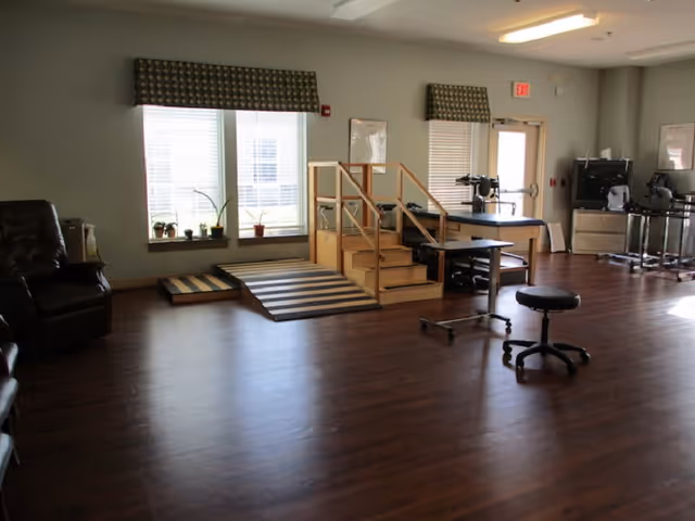A spacious interior therapy room with hardwood floors, mobility ramps and steps, medical equipment, stools and seating near windows.
