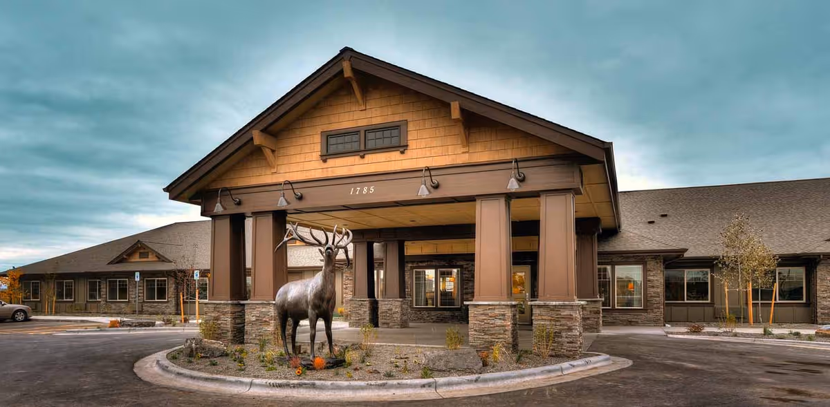 Front exterior of a memory care community showing a covered entrance with columns and a large elk statue in the roundabout.