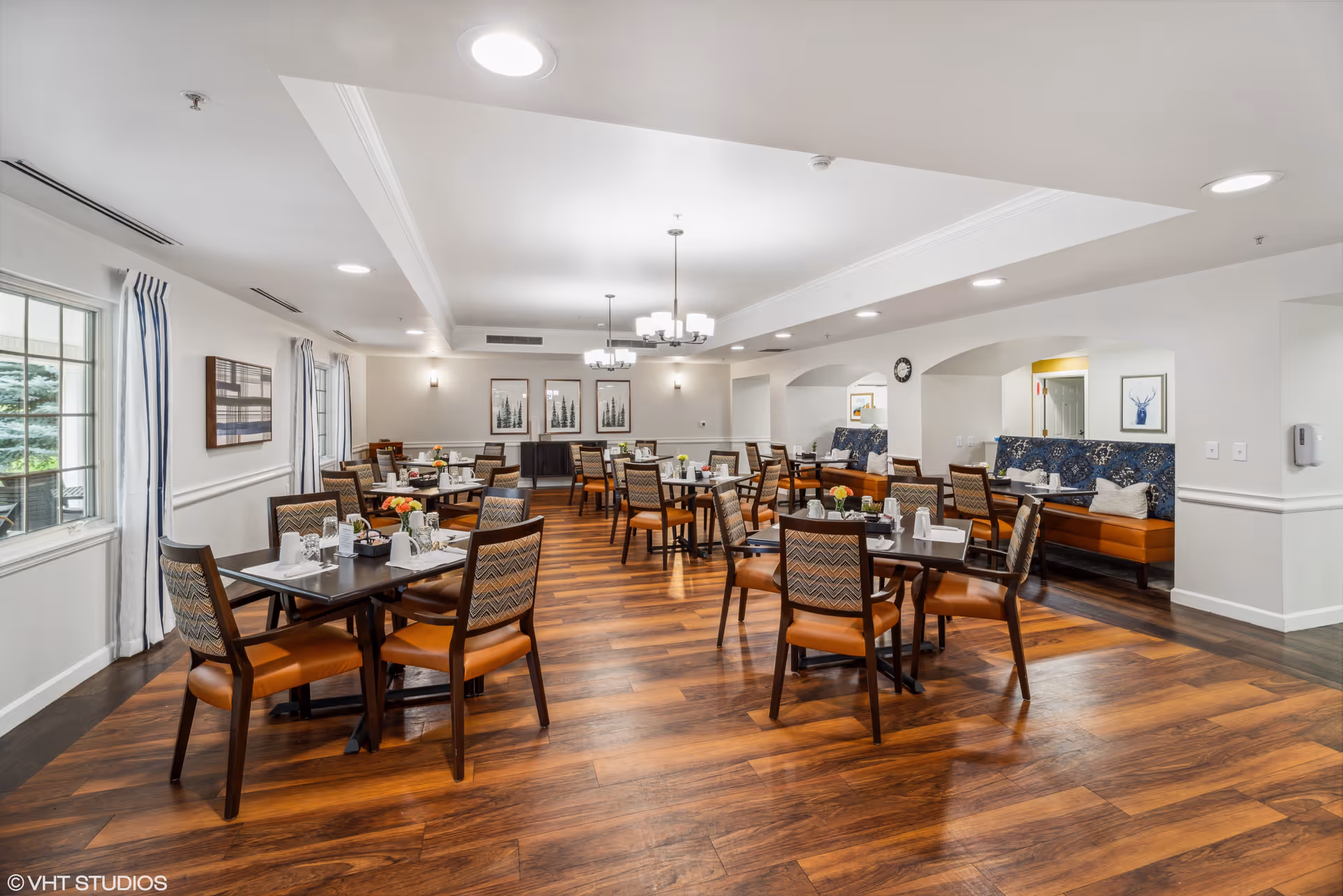 A spacious dining room with multiple tables and chairs arranged neatly. The room features wooden flooring, white walls, and large windows with curtains allowing natural light to enter. There are decorative framed pictures on the walls and a few flower arrangements on the tables. The ceiling has recessed lighting and chandeliers. A seating area with cushioned benches is visible in the background.