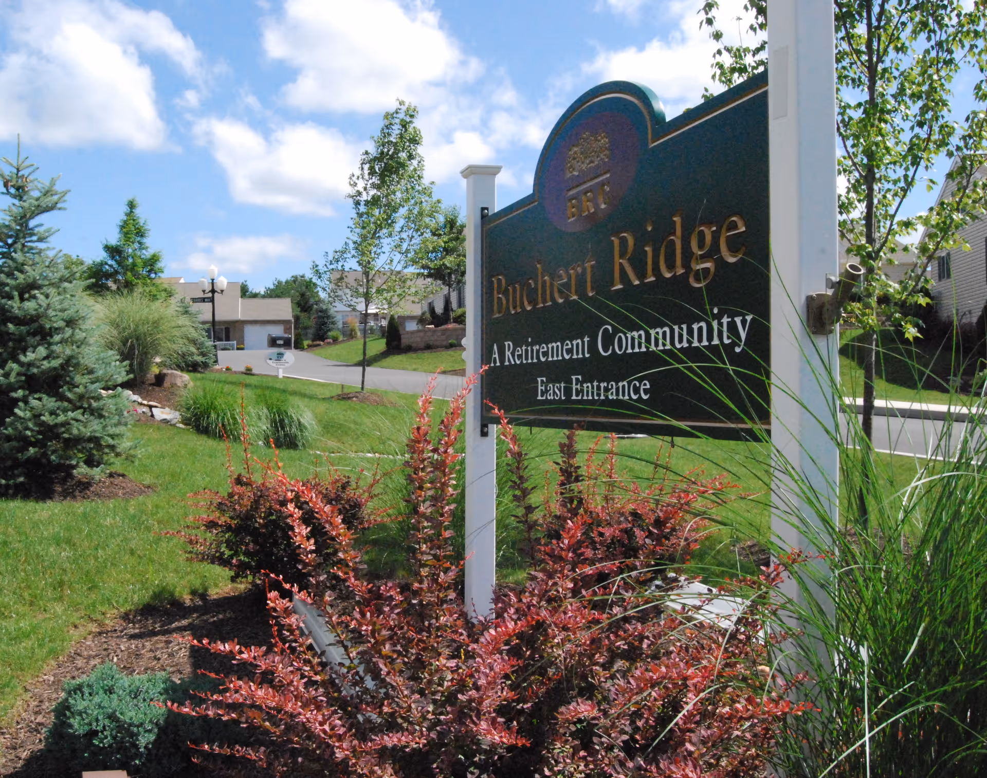 Outdoor view of the entrance sign for Buchert Ridge, a retirement community, surrounded by landscaped greenery including bushes, grass, and trees under a partly cloudy blue sky.