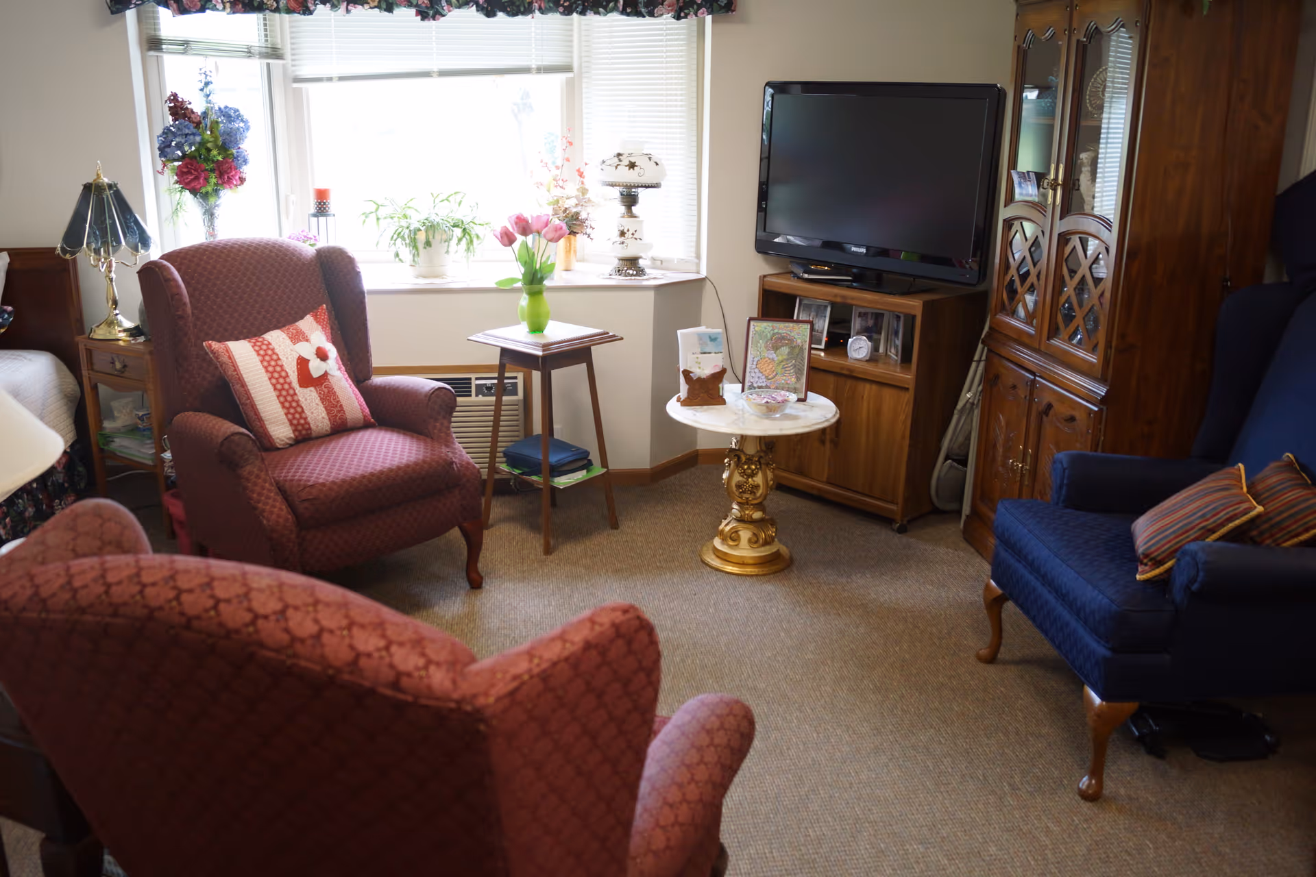 A cozy living room area with two red patterned armchairs and one blue armchair with striped pillows. There is a small round table with a decorative base holding framed pictures and a bowl, a wooden cabinet with a flat-screen TV on top, and a window with floral curtains letting in natural light. Various plants and flowers are placed on a small table and windowsill.