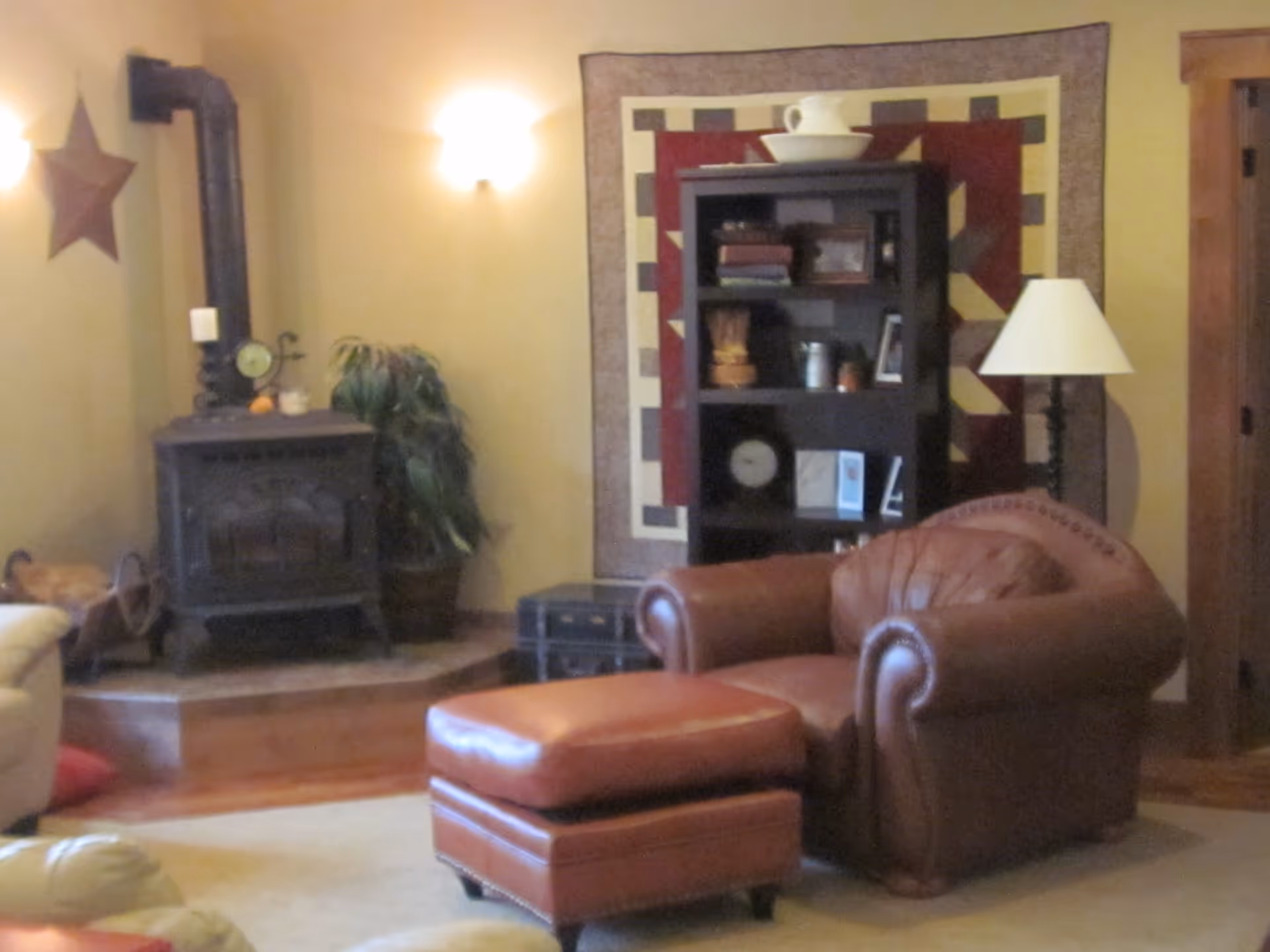 Cozy living room with a brown leather armchair and matching ottoman in the foreground. Behind the chair is a black bookshelf with various decorative items and books, set against a wall with a quilt hanging. To the left, there is a wood-burning stove with a pipe extending upward, a potted plant, and a star decoration on the wall. A floor lamp with a white shade stands to the right of the bookshelf.