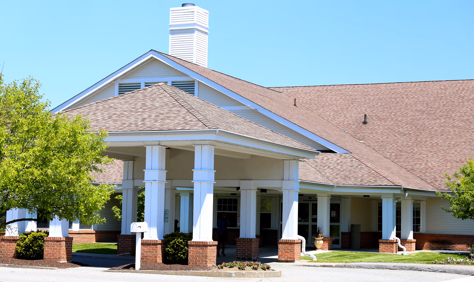 Exterior view of a senior living facility building with a covered entrance supported by white columns with brick bases, surrounded by green trees and landscaping under a clear blue sky.