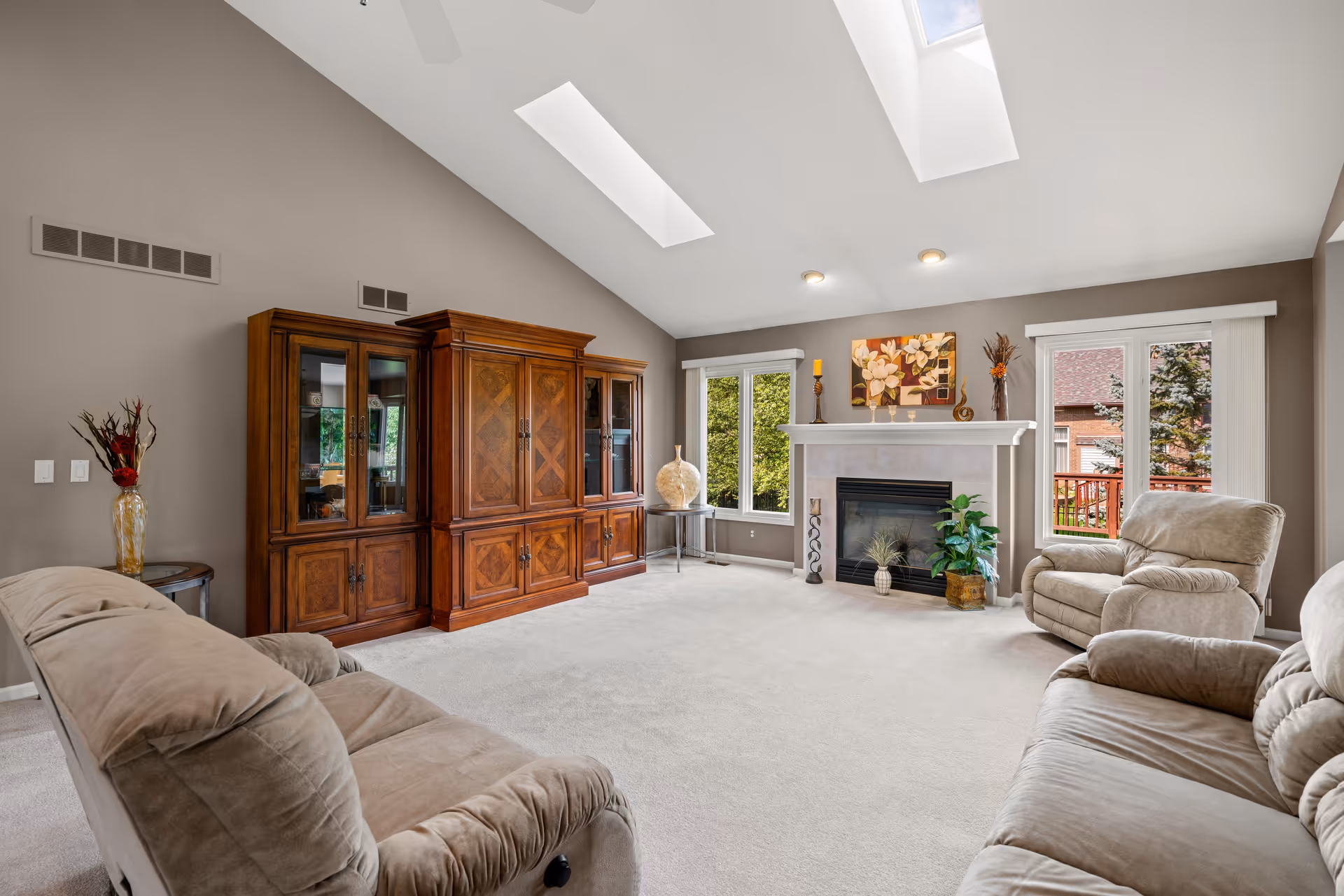 A spacious living room with beige carpet and taupe walls, featuring two skylights in the vaulted ceiling. The room contains a large wooden entertainment cabinet, a fireplace with decorative items on the mantel, two beige recliner chairs, and a beige sofa. There are large windows and a sliding glass door that provide natural light and a view of greenery outside.