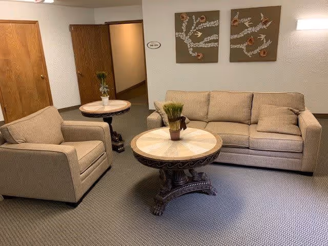 A cozy living room area with a beige sofa and matching armchair, two round wooden tables with decorative plants on top, beige carpeted floor, wooden doors, and two wall art pieces featuring floral designs on a light-colored wall.
