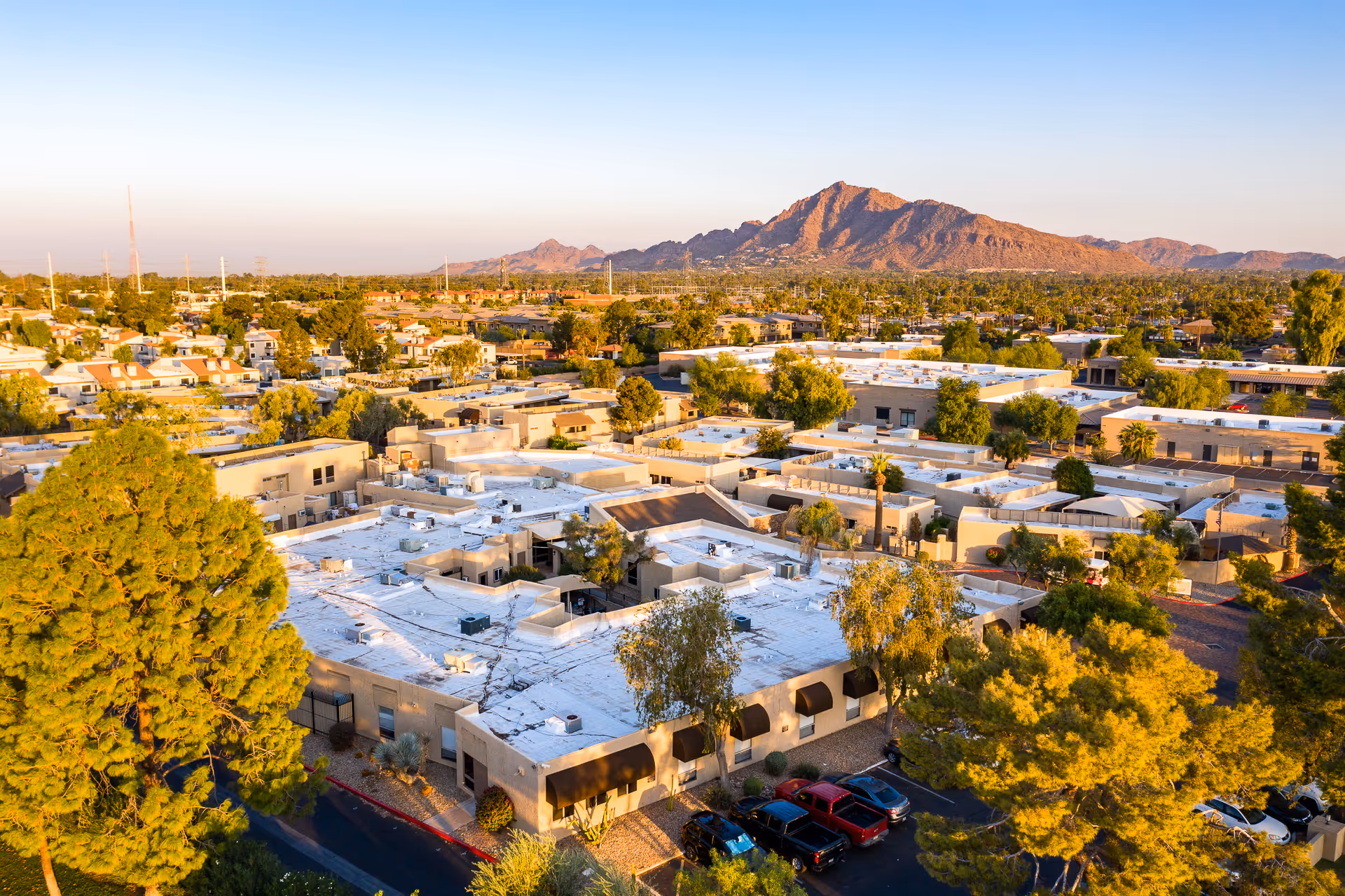 Aerial view of Scottsdale Village Square senior living facility with multiple beige buildings featuring flat white roofs, surrounded by trees and parked cars, with a mountain range in the background under a clear sky.