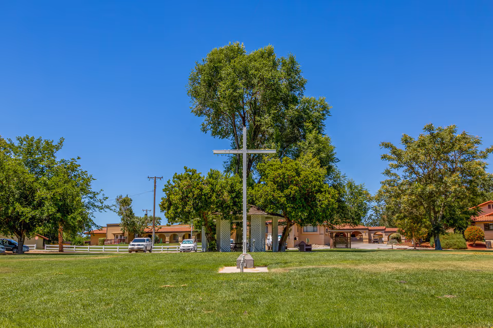 A large green lawn with a tall metal cross in the center, surrounded by trees and residential buildings under a clear blue sky.