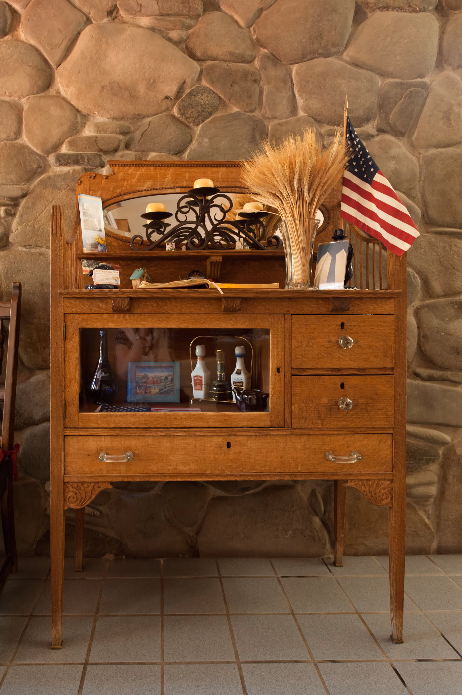 An antique wooden sideboard with glass-fronted cabinet and drawers, decorated with a metal candle holder with candles, a vase of dried wheat stalks, and a small American flag, set against a stone wall background.