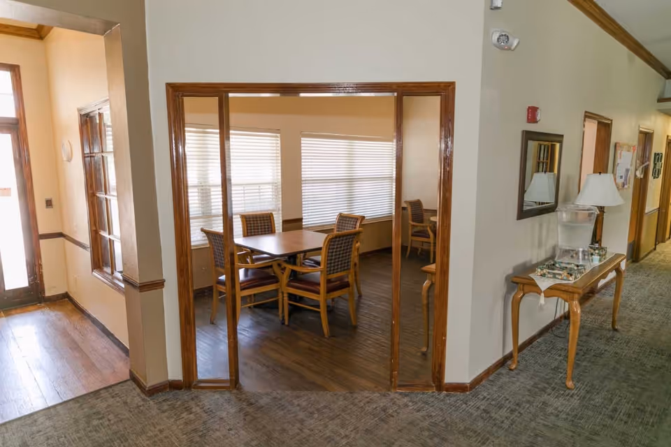 Interior view of a senior living facility showing a hallway with carpeted floor and a wooden table with a lamp and water dispenser. To the left, there is a room with wooden flooring and a table surrounded by chairs, illuminated by natural light from windows with blinds.