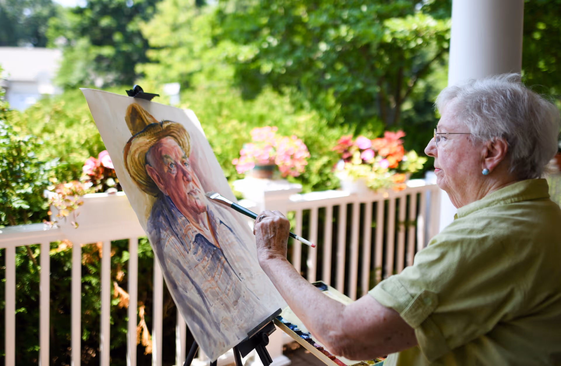 An elderly woman with short gray hair and glasses is painting a portrait of an older man wearing a straw hat on an easel outdoors. She is seated on a porch with white railing, surrounded by green trees and colorful flowers in the background.