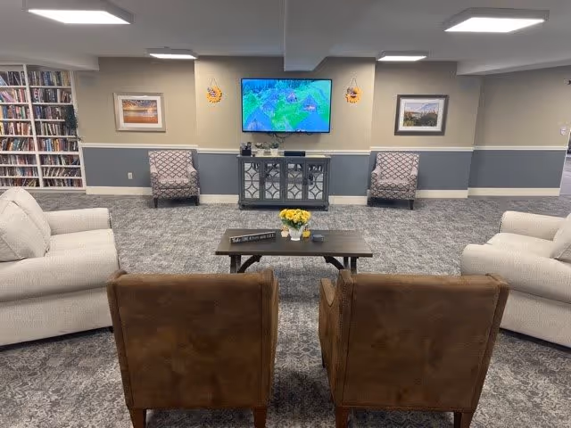 A cozy senior living common area with two brown armchairs facing a coffee table with a vase of yellow flowers, flanked by two white sofas. In the background, there are two patterned armchairs, a TV mounted on the wall above a cabinet, framed pictures on the wall, and a bookshelf filled with books on the left side.
