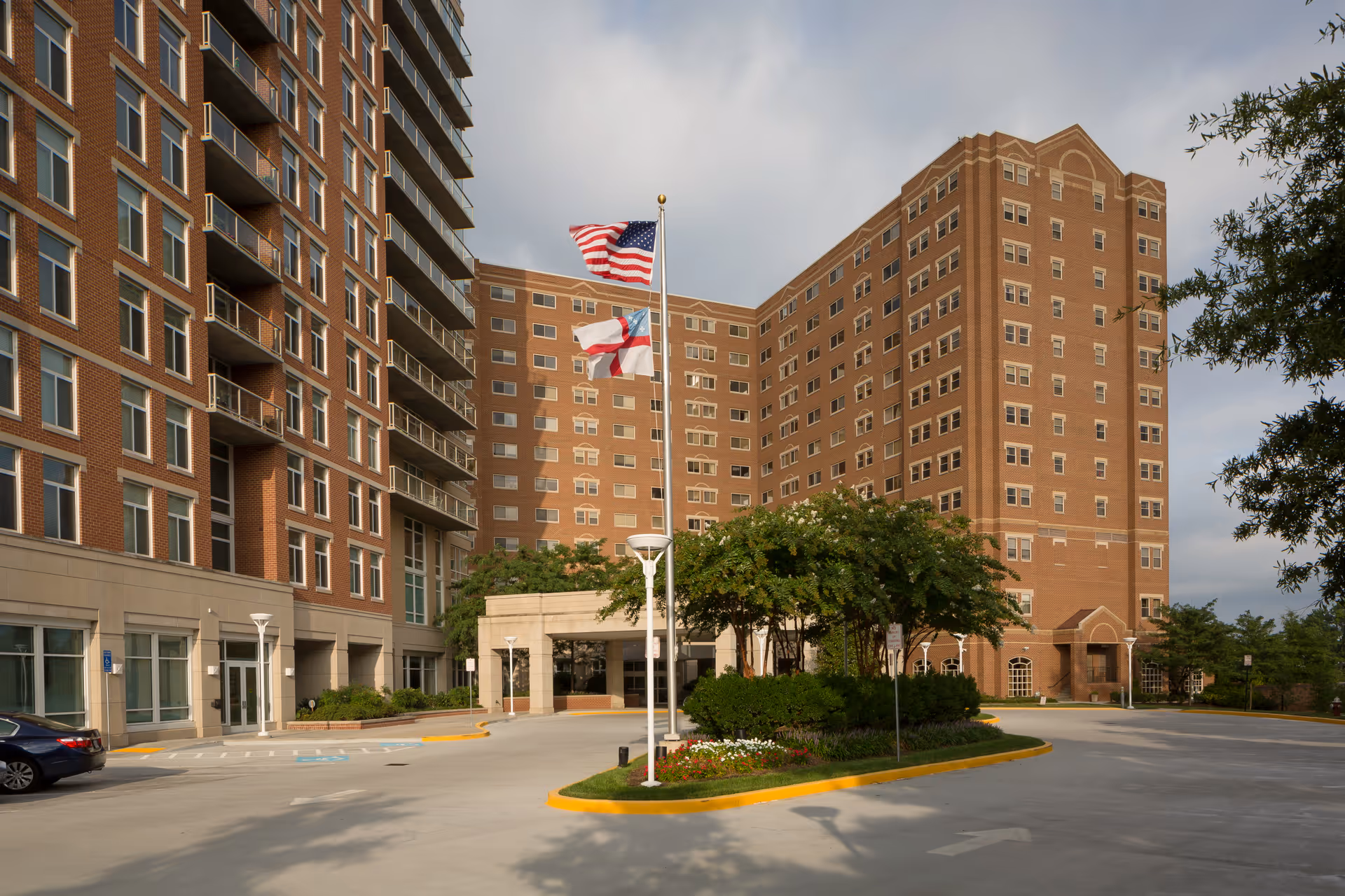Exterior view of a large multi-story brick building with many windows, featuring a driveway with a landscaped island in the center. Two flagpoles display the American flag and another flag. Trees and shrubs surround the entrance area under a partly cloudy sky.