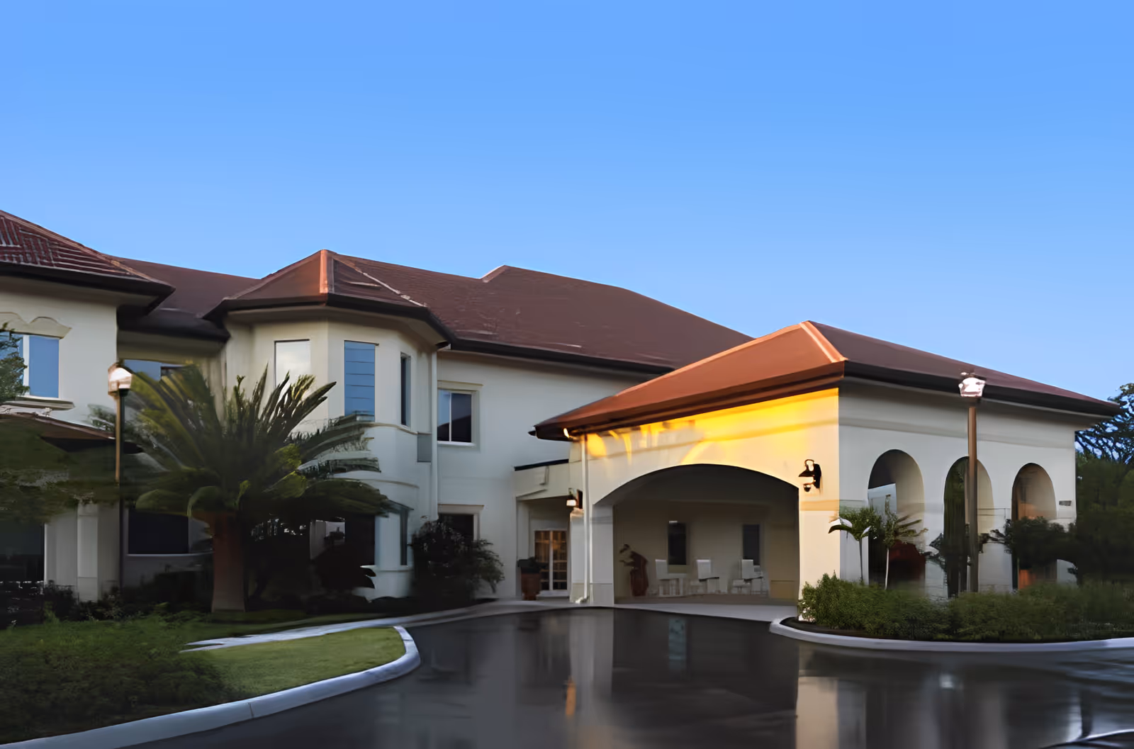 Exterior view of a senior living facility building with a covered entrance, arched openings, and a red roof under a clear blue sky. There are palm trees and landscaped greenery around the driveway.