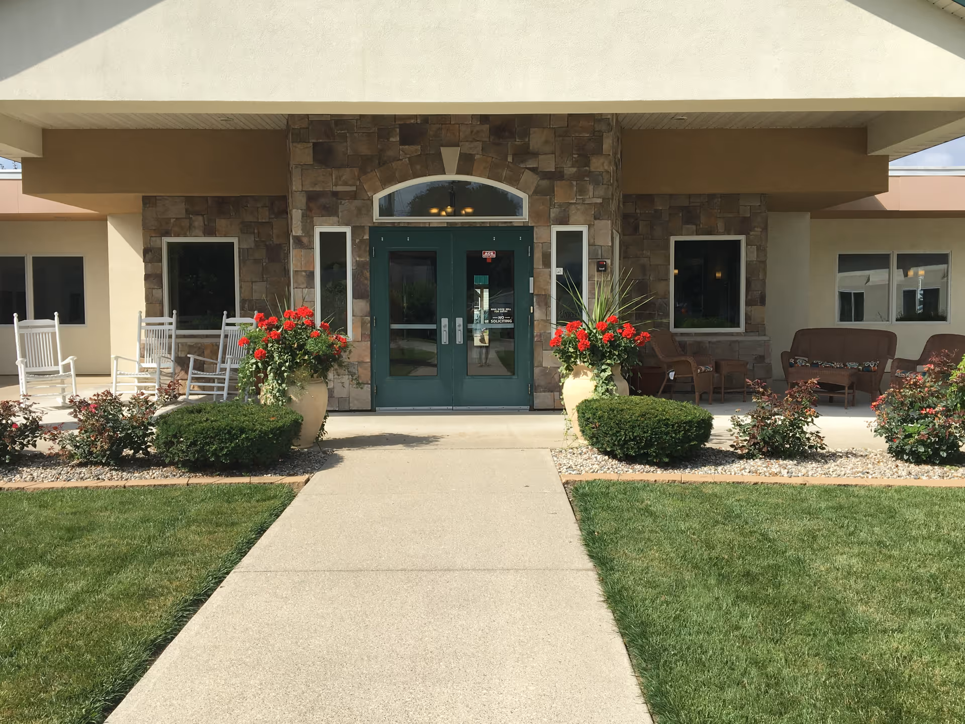 Concrete walkway leading to the front entrance of an assisted living building with double glass doors, potted red flowers, rocking chairs and patio seating.