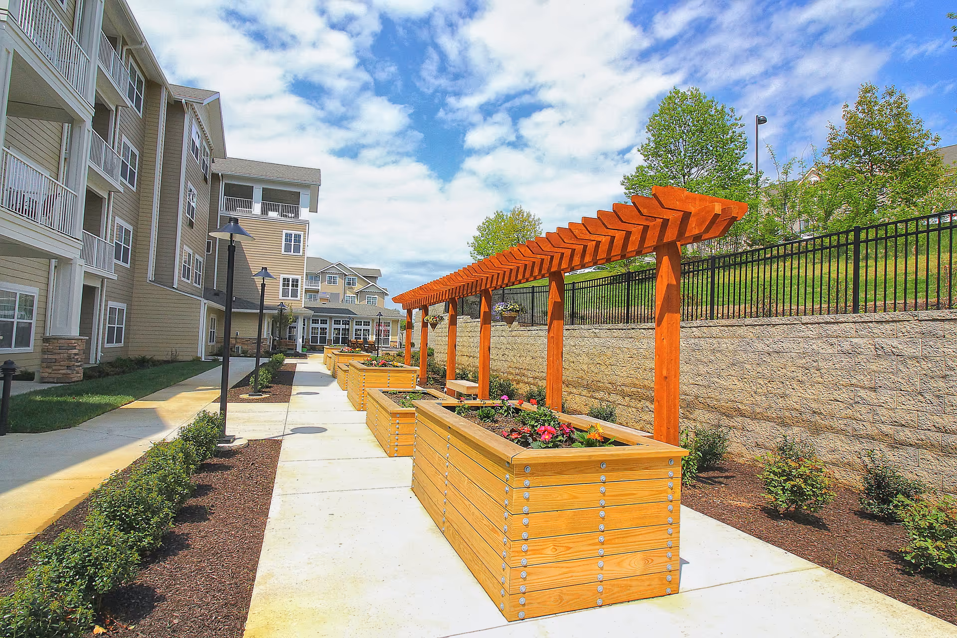 Outdoor walkway at Ivy Creek Gracious Retirement Living featuring wooden planter boxes with flowers and a wooden pergola structure. The path is lined with small bushes and lamp posts, with a multi-story residential building on the left and a stone retaining wall with trees on the right under a partly cloudy sky.