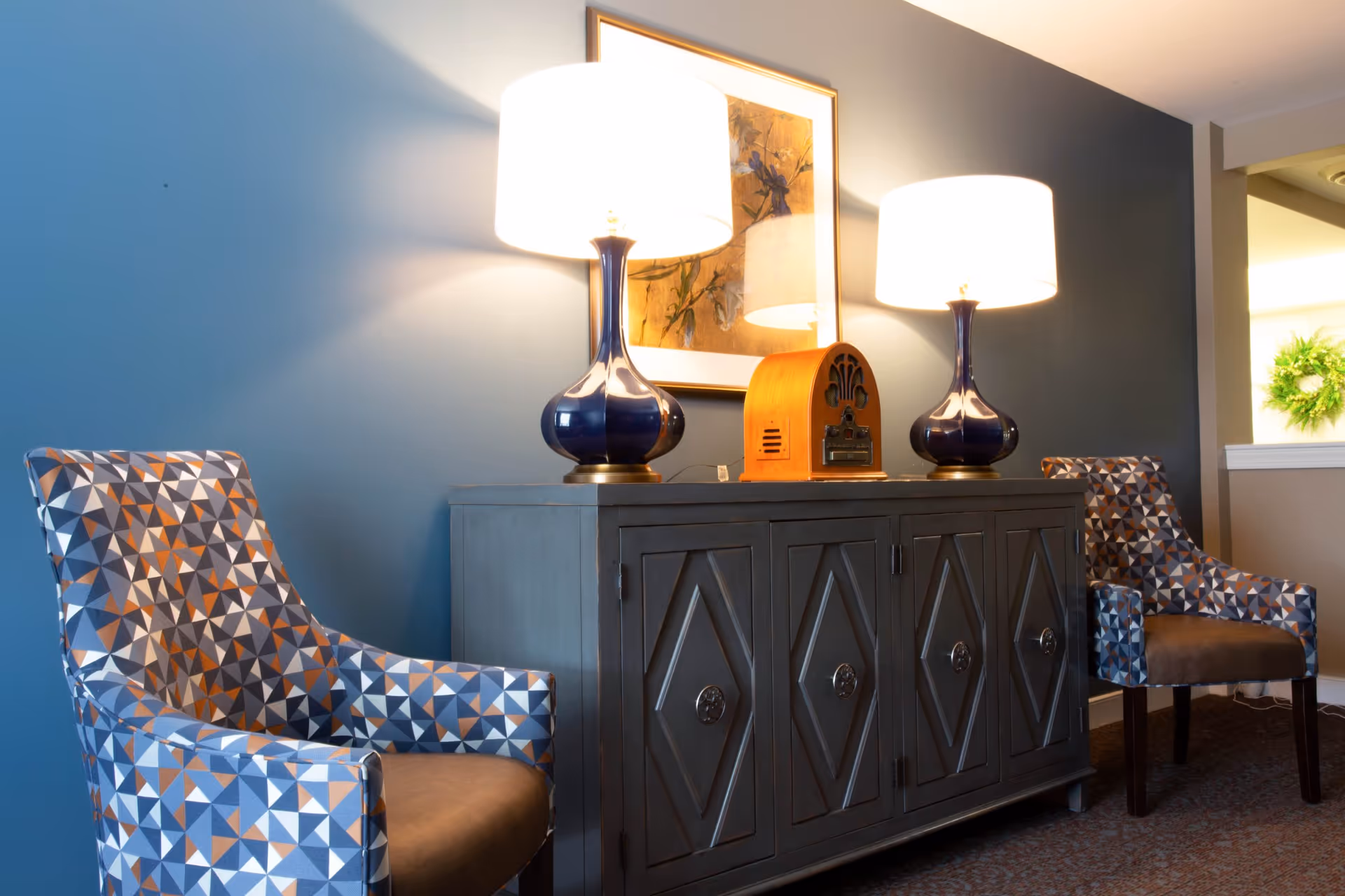 Two patterned armchairs flank a dark wooden cabinet with diamond-shaped designs on the doors. On top of the cabinet are two blue table lamps with white shades and a vintage-style wooden radio. A framed floral artwork hangs on the blue wall behind the cabinet.