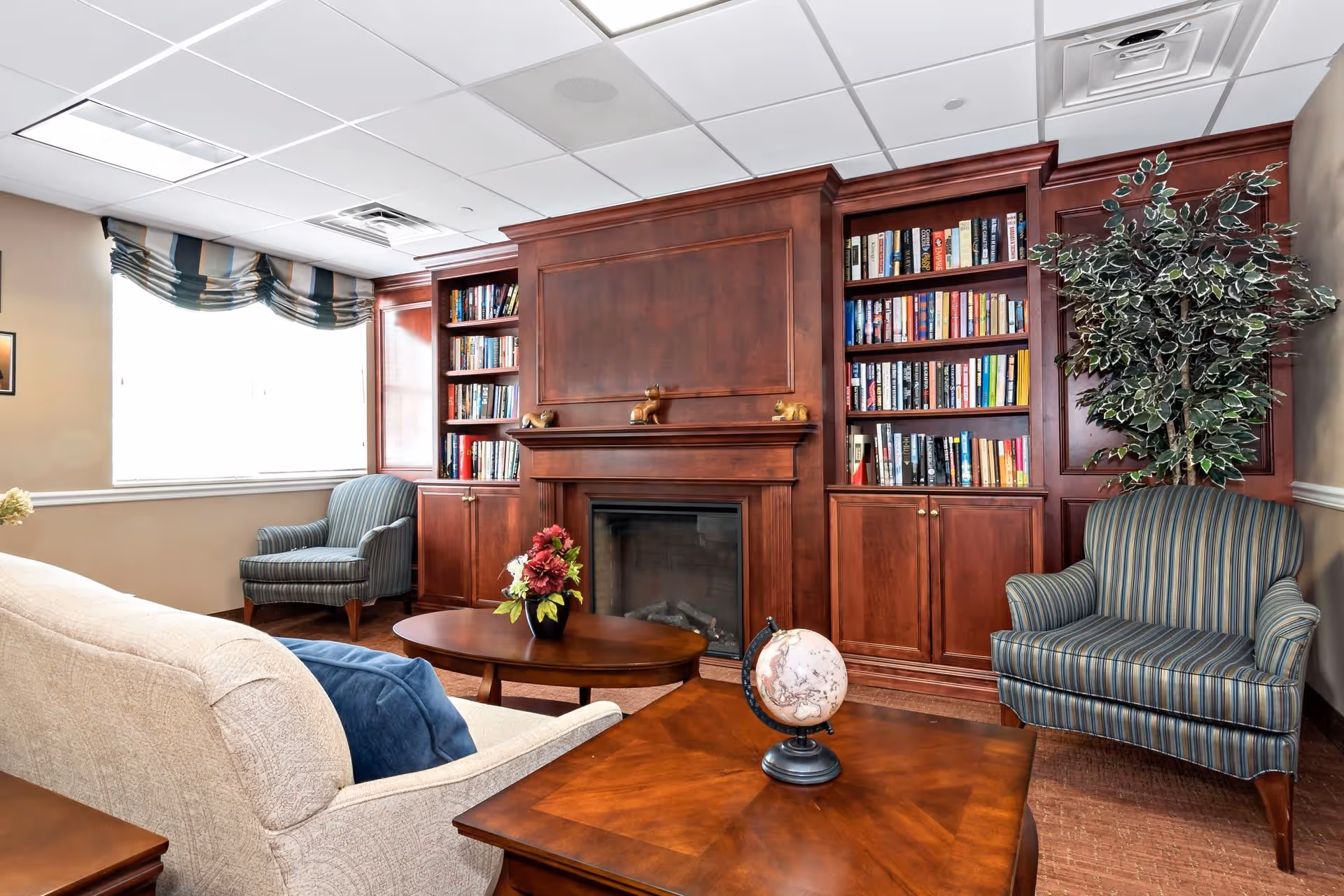 A cozy living room area featuring a beige sofa with a blue pillow, two striped armchairs, a wooden coffee table with a globe and a smaller table with a flower arrangement. The background has built-in wooden bookshelves filled with books and a fireplace in the center. A large window with striped curtains allows natural light into the room.