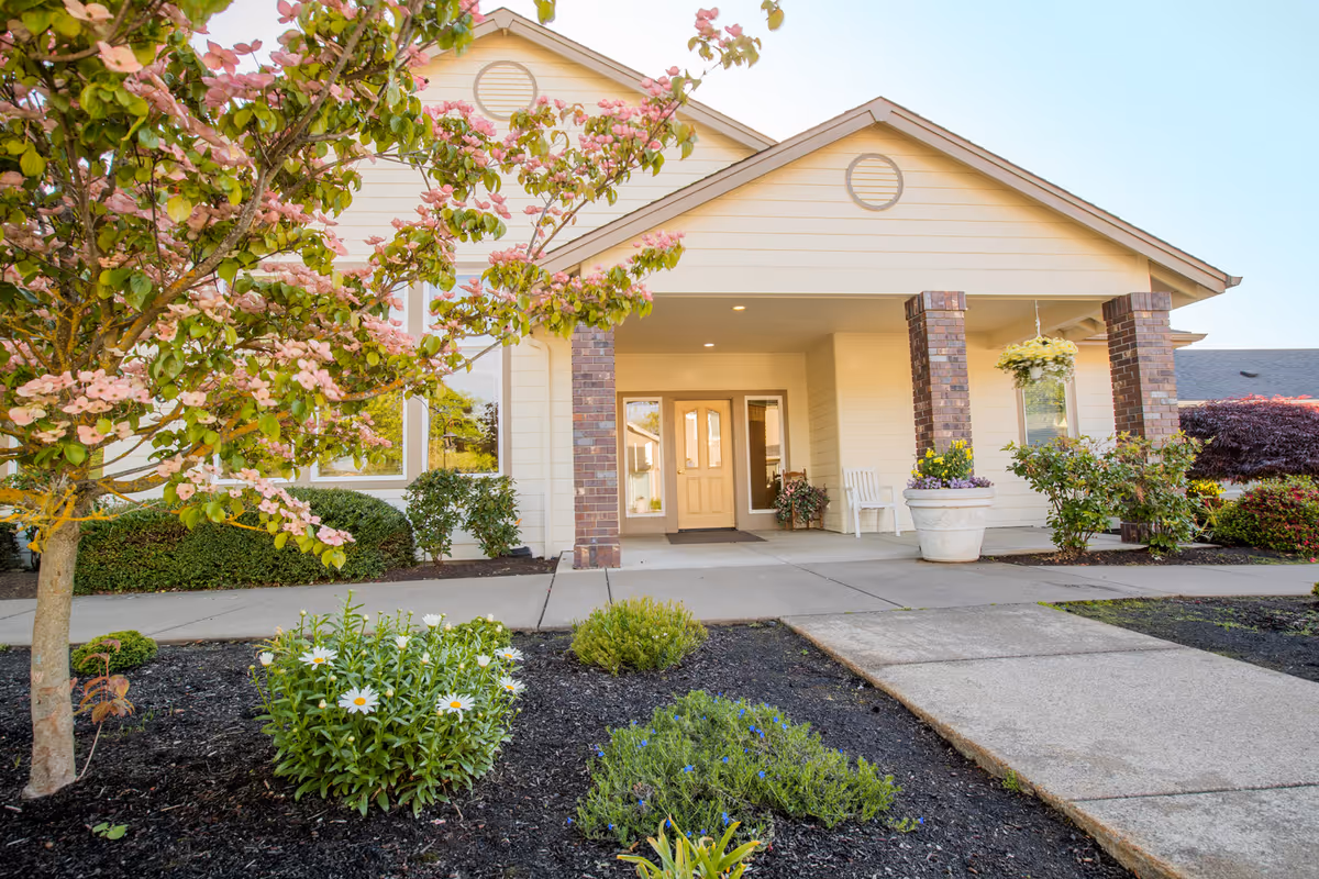 Front exterior view of a single-story building with a covered entrance supported by brick columns. There is a concrete walkway leading to the double front doors. The landscaping includes a flowering tree with pink blossoms, green shrubs, and white daisies in the garden beds.