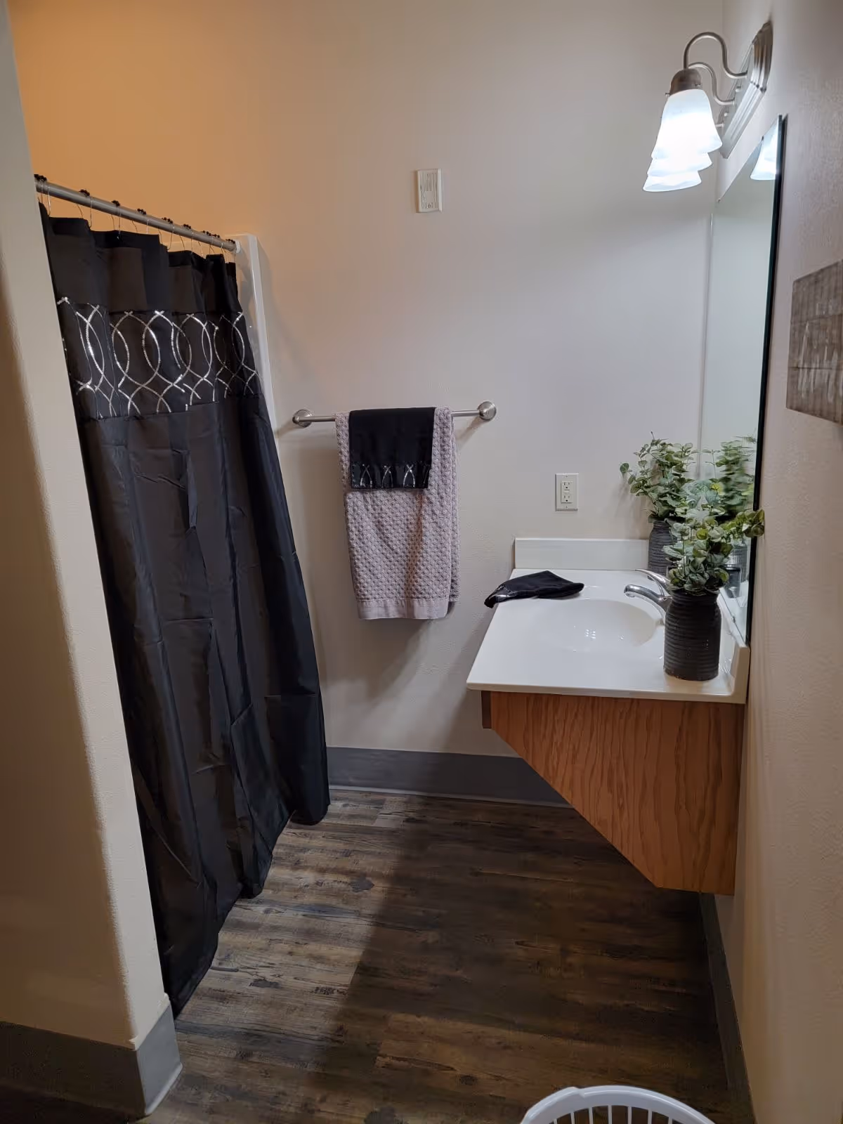 A bathroom with a dark shower curtain featuring a circular pattern, a towel rack with two towels hanging, a white sink countertop with a faucet, a mirror above the sink, a potted plant on the countertop, and wall-mounted light fixtures above the mirror. The floor has a wood-like finish.