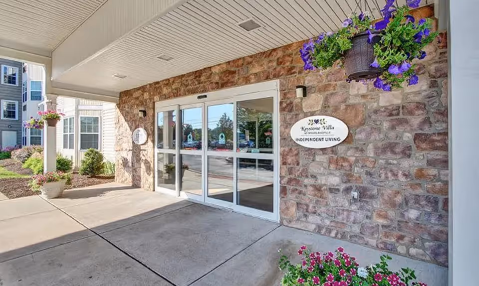 Entrance to Keystone Villa at Douglassville featuring automatic sliding glass doors set in a stone wall exterior. There are hanging flower pots with purple flowers and planters with pink flowers near the entrance. A sign on the wall reads 'Keystone Villa Douglassville Independent Living.'