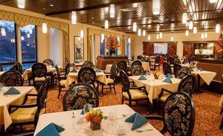A spacious dining room with multiple tables covered in white tablecloths and set with blue folded napkins, glassware, and floral centerpieces. The room features patterned upholstered chairs, large windows with curtains, warm lighting from ceiling fixtures, and decorative elements including framed artwork and flower arrangements.