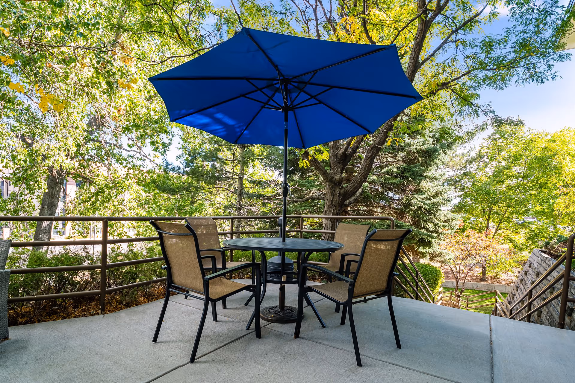 Outdoor patio area with a round table, four chairs, and a large blue umbrella providing shade. The patio is surrounded by trees and greenery, with a metal railing and stairs leading down to a garden area.