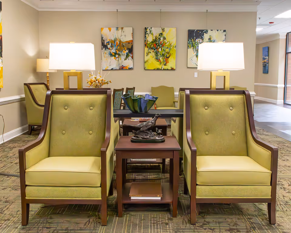 A well-lit lounge area with two green upholstered armchairs facing forward, separated by a wooden side table with a decorative bird sculpture. Behind the chairs is a long table with a blue and green glass bowl and two modern lamps with white shades. The walls are adorned with three abstract paintings in yellow, green, and orange tones. The room has a carpeted floor and a hallway visible to the right.