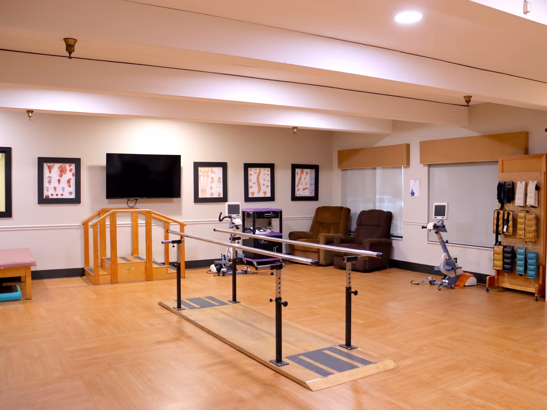 A rehabilitation therapy room with parallel walking bars, exercise equipment, recliners, and a wall-mounted TV.