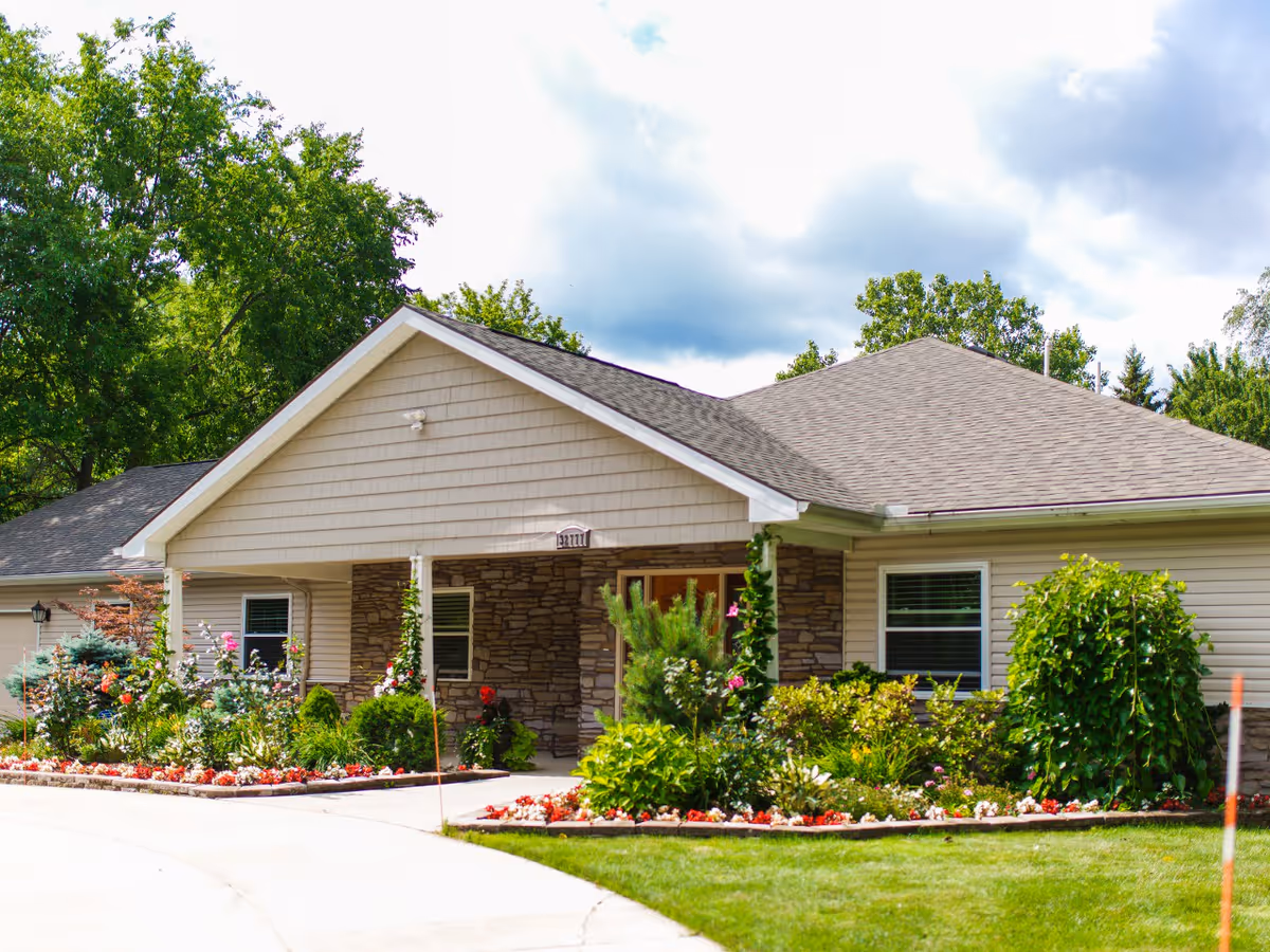Single-story building with beige siding and stone accents, surrounded by well-maintained flower beds and greenery under a partly cloudy sky.