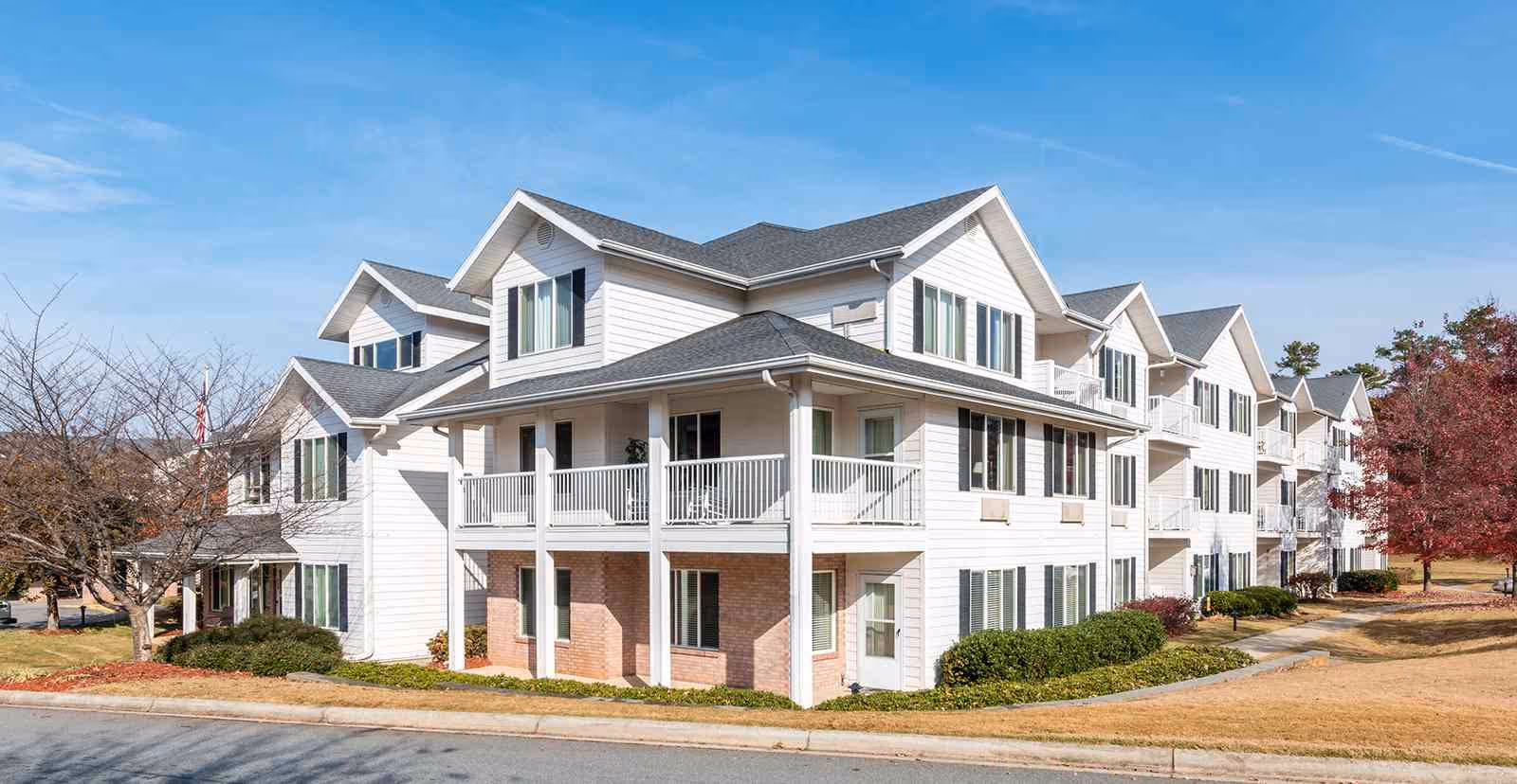 Three-story white senior living building with balconies, gabled roofs, and surrounding landscaping under a blue sky.