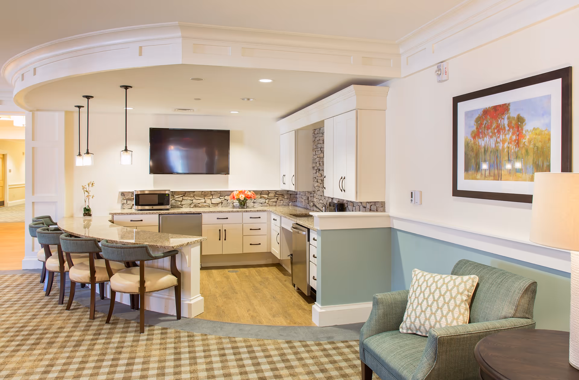 A modern kitchen area in a senior living facility with a curved granite countertop and five cushioned bar stools. The kitchen features white cabinets, a stone backsplash, a microwave, a dishwasher, and a wall-mounted flat-screen TV. Adjacent to the kitchen is a comfortable green armchair with a patterned pillow, a side table, and a lamp. A framed painting of trees with autumn colors hangs on the wall.