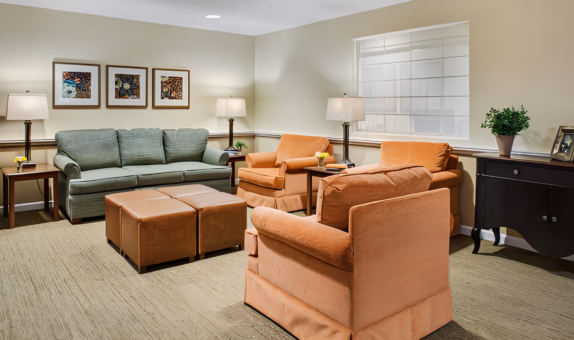 A cozy living room with a green sofa, two orange armchairs, and two brown ottomans. The room is softly lit by three table lamps placed on side tables. There are three framed artworks on the wall above the sofa, a window with blinds, a dark wooden sideboard with a potted plant and picture frames, and a beige carpet covering the floor.