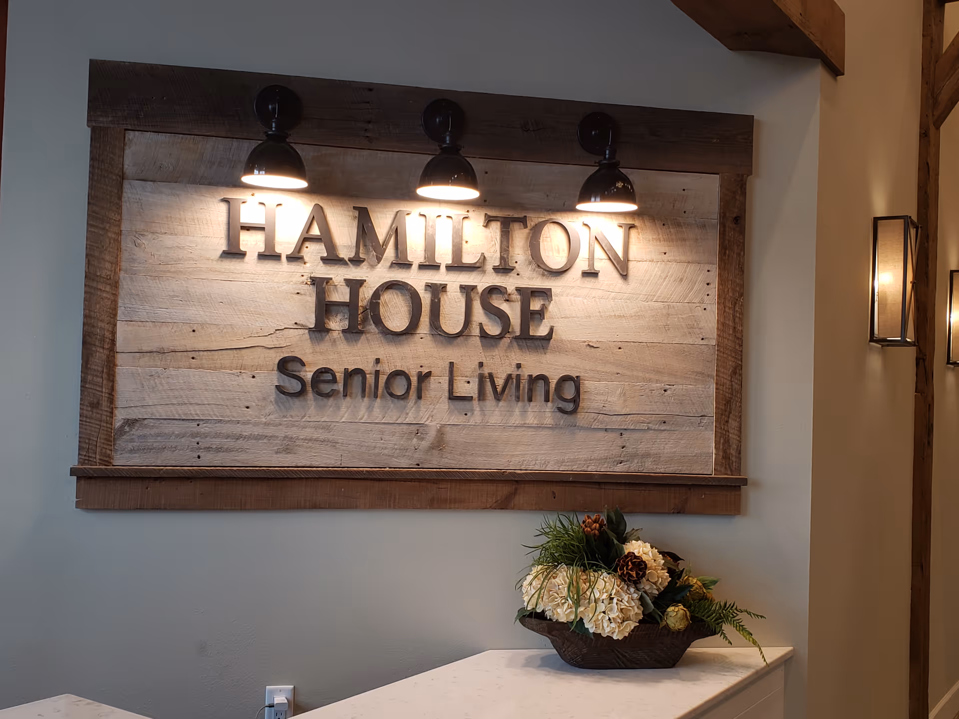 Wooden wall sign reading "Hamilton House Senior Living" mounted above a table with a floral arrangement under three lamps.