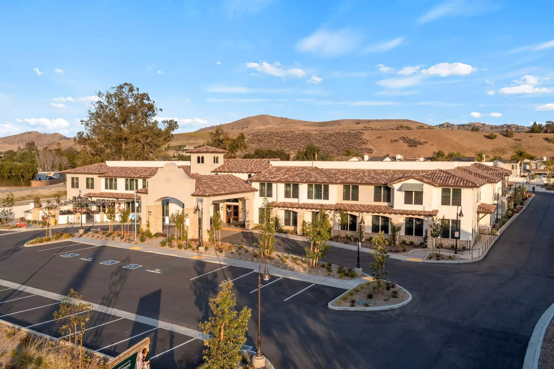 Front exterior aerial view of a two-story Mediterranean-style senior living building with a parking lot and hills in the background.