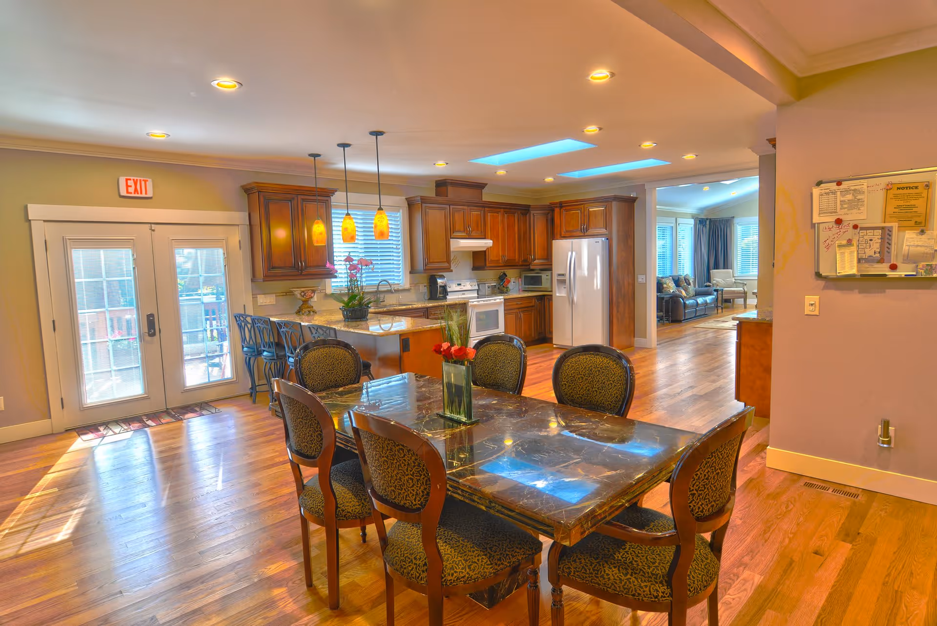 A spacious and well-lit kitchen and dining area with wooden floors and cabinetry. The dining table has a dark marble top with six upholstered chairs around it. The kitchen features modern appliances including a refrigerator, stove, and microwave. Pendant lights hang above the kitchen island with bar stools. In the background, a living room area with sofas and large windows is visible. A bulletin board with notices is mounted on the wall to the right.