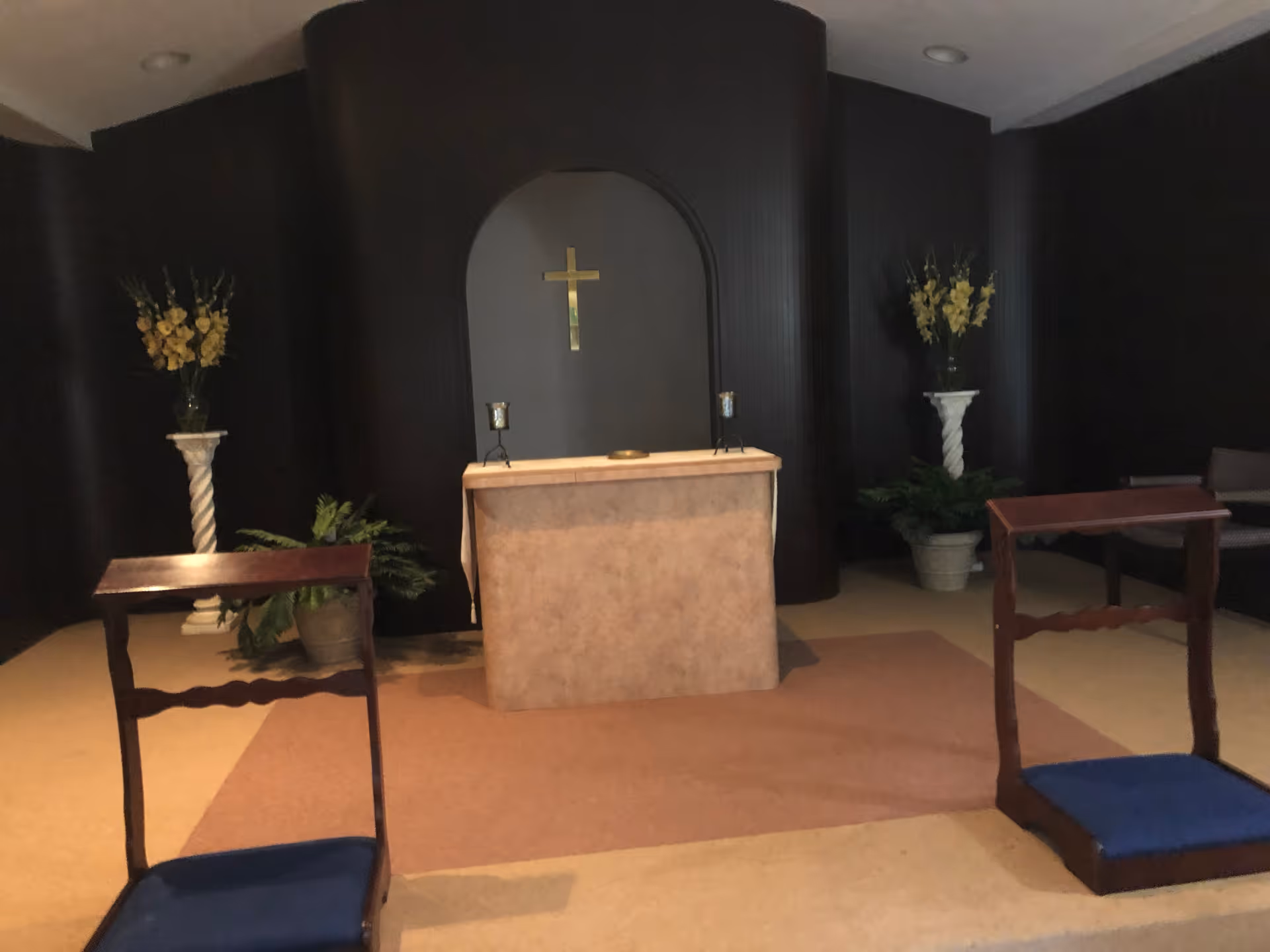 Interior of a small chapel or prayer room with a central altar covered in a beige cloth, a gold cross mounted on a dark wood-paneled wall behind it, two candle holders on the altar, two kneeling benches with blue cushions in the foreground, and floral arrangements on white pedestals on either side of the altar.